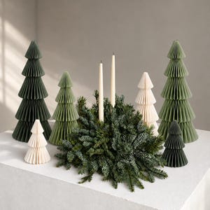 Puede incluir: Una colección de árboles de Navidad de papel de nido de abeja en tonos verde oscuro, verde salvia y crema, dispuestos alrededor de una corona de abeto central con dos velas cónicas altas, delgadas y de color crema en el centro.