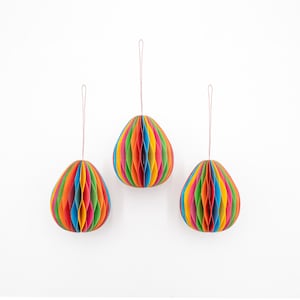 Puede incluir: Tres adornos de papel de panal de abeja coloridos que cuelgan de cuerdas. Cada adorno es una combinación de colores diferente de rojo, naranja, amarillo, verde, azul y morado.