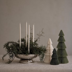 May include: A silver pedestal bowl filled with dark green evergreen branches and four tall white taper candles, accompanied by three paper honeycomb Christmas trees in varying sizes and shades of cream and muted green.