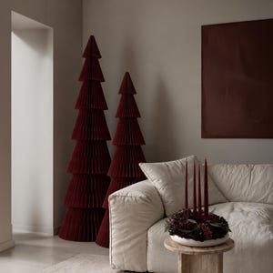 Op de afbeelding: Twee hoge, donkerrode, geplooide papieren kerstbomen staan naast een beige bank. Op een kleine houten tafel voor de bank staat een witte schaal gevuld met donkerrode bloemen en vier hoge, slanke, donkerrode dinerkaarsen.