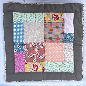 May include: A square patchwork quilt cover featuring a central grid of various fabric squares in floral, polka dot, and geometric patterns. The centre is framed by a solid dark grey border and finished with a thin, patterned fabric edge trim.