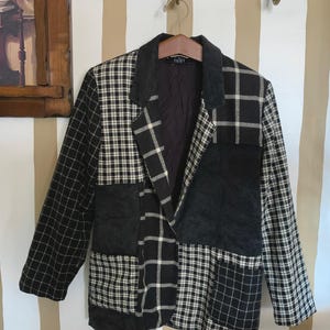 May include: A patchwork blazer in black and white, featuring a mix of plaid and corduroy panels. The jacket has a notched lapel and is hanging on a wooden hanger. The lining is visible, and the overall style is vintage.