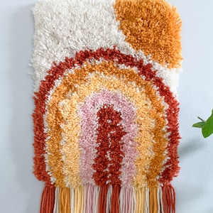 May include: A textured wall hanging with a rainbow design in shades of orange, pink, and yellow. The top has a white and orange textured background. The bottom has a fringe of matching colours. A unique home decor item.