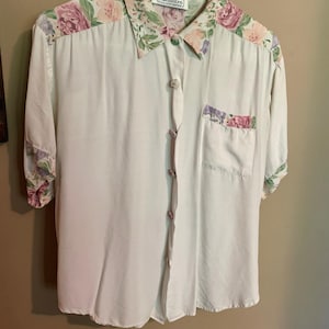 May include: A cream-colored button-down shirt with a floral collar, cuffs, and pocket detail. The floral pattern includes pink and purple roses and green leaves. The shirt has the word "impressions" on the collar.