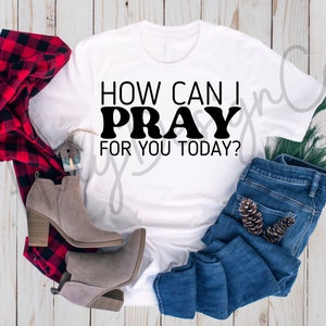 Puede incluir: Camiseta blanca con texto negro que dice "How Can I Pray For You Today?" La camiseta está colocada sobre una superficie de madera con una camisa de cuadros rojos y negros, un par de botas marrones, unos vaqueros azules y una rama de pino.