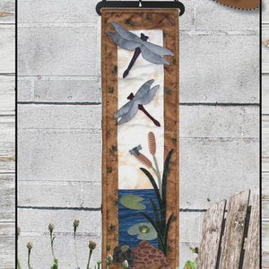 May include: A fabric wall hanging featuring a pond scene with dragonflies, cattails, and a turtle. The banner is 6 inches wide and 21 inches long. The text "Pond Critters" is at the top, and the skill level is "EASY".
