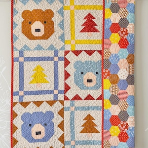 May include: A handmade quilt featuring bear and tree designs in various colors. The quilt has a red border and is hung with wooden clothespins. The design includes brown, blue, yellow, and red elements, with a section of colorful hexagonal patterns.