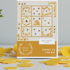 May include: A quilt pattern titled "Honey to the Bee" by Sue Daley and co. The design features a grid of honeycomb and bee illustrations. The packaging is white and gold, with yellow honeycomb-shaped confetti scattered around.