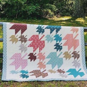 May include: A handmade quilt featuring a leaf pattern in shades of red, pink, blue, and brown on a cream background. The quilt is displayed outdoors, draped over a wooden fence, with a green forest backdrop.