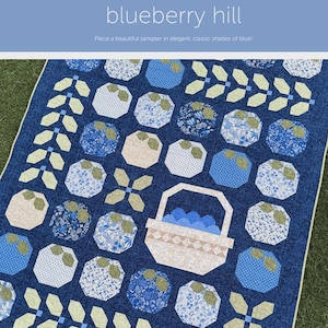 May include: A blue quilt with a repeating pattern of octagons and floral designs. The quilt features a basket filled with blue objects. The text "blueberry hill" is at the top, with the designer's name at the bottom.