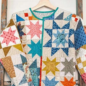 May include: A colorful, quilted jacket pattern titled "Beachcomber" by Laundry Basket Quilts. The jacket features a star pattern in various colors, including blue, pink, orange, and yellow. The jacket is hanging on a wooden hanger.