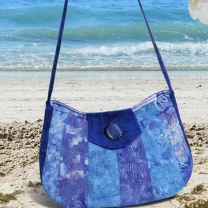 May include: A blue and purple Catalina Sling bag with a floral pattern, set against a beach backdrop. The bag has a curved shape and a flap closure. Text on the image reads "Pink Sand Beach Designs" and "Catalina Sling".