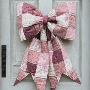 May include: A large, quilted bow in shades of pink and burgundy hangs on a white door. The bow is made of patchwork squares and has long, pointed tails. Text at the top reads "THE BIG GIANT QUILTED BOW".