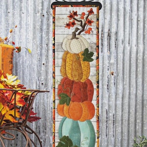 May include: A vertical fall-themed wall hanging featuring a stack of colorful pumpkins. The finished size is 6" x 21.5". The design includes a variety of orange, yellow, and green pumpkins with leaf accents. The text "Pumpkins Galore" is displayed at the top.