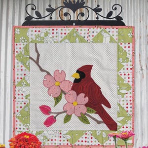 May include: A quilted wall hanging featuring a cardinal bird perched on a flowering branch. The artwork is framed with a pink border and green floral accents. The text "CARDINAL BLOOMS" and dimensions 16.5" x 16.5" are displayed.