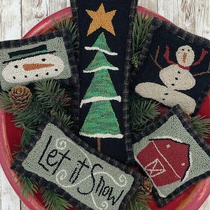 May include: A collection of five punchneedle bowl fillers with winter holiday themes. Designs include a snowman, Christmas tree, barn, and the text "Let it Snow." The fillers are arranged in a red bowl with pine needles.