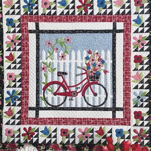 May include: A quilted wall hanging featuring a red bicycle with a basket of flowers in front of a white picket fence. The quilt has a floral and geometric border, with the text "Joy Ride" at the top. Finished size is 44 x 44 inches.