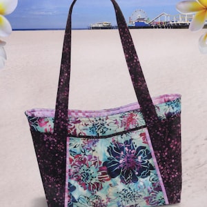 May include: A tote bag with a floral pattern and the text "Pink Sand Beach Designs" and "Brentwood Bag". The bag has shoulder straps, a zipper top, and multiple pockets. The bag's design includes a floral pattern with purple, blue, and pink colors.