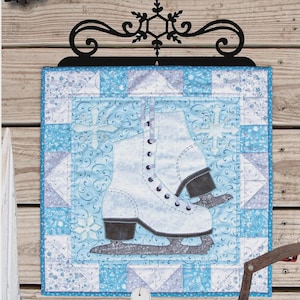 May include: A quilted wall hanging featuring a white ice skate design on a blue and white patterned background. The text "i-SKATE" is displayed above the image, with dimensions of 16.5" x 16.5".
