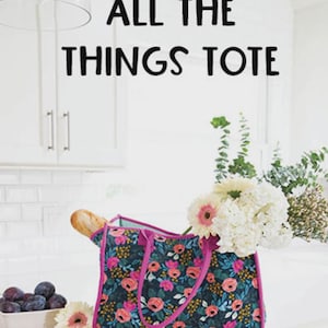 May include: A rectangular fabric tote bag featuring a dark navy blue background with a vibrant floral pattern of pink, orange, and yellow flowers and green leaves. The bag has bright pink trim and handles, with the text ALL THE THINGS TOTE above it.