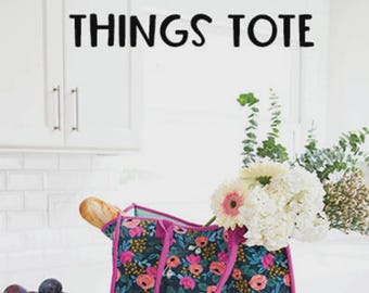 May include: A floral tote bag with pink trim, filled with flowers and a baguette. The bag is on a wooden cutting board. Text on the bag reads "ALL THE THINGS TOTE". The brand logo "KNOT + THREAD design" is visible.