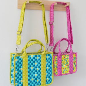 May include: Two crossbody tote bags hanging on a wooden rack. The larger bag has a blue and yellow checkered pattern, while the smaller bag has a floral design. The bags have matching straps and handles. Text at the top reads "Zuri Crossbody Tote".