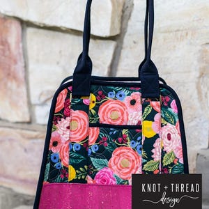 May include: A floral print carryall bag with a dark navy blue trim and handles. The bag features a vibrant pattern of pink, red, and yellow flowers on a dark background. The bottom panel is a solid pink. The text "Boundless Carryall" is at the top.