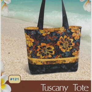 May include: Tuscany Tote bag with a floral pattern in orange and yellow on a navy blue background. The tote has black handles and a yellow and black striped band. The background shows a beach scene with the text "Pink Sand Beach Designs".