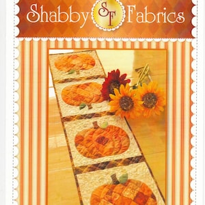 K&ouml;nnte beinhalten: Dekorativer Tischl&auml;ufer mit Patchwork-K&uuml;rbismotiven. Der L&auml;ufer hat einen cremefarbenen Hintergrund mit orangefarbenen und braunen K&uuml;rbisapplikationen. Der Text "Shabby Fabrics" und "Patchwork Pumpkin Table Runner" sind sichtbar.