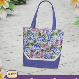 May include: A tote bag with a floral pattern of blue and pink hydrangeas. The bag has a blue base and handles, with the text "Capri Carryall" and "6 POCKETS INSIDE" on the bottom. The bag is approximately 14 inches tall.