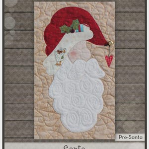 May include: A fabric quilt block titled The Whole Country Caboodle Pre-Santa featuring a Santa Claus applique with a red hat, white beard, and holly accents on a tan background. Includes precut fused applique for 8 inch by 14 inch block.