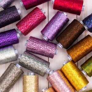 May include: A collection of spools of metallic thread in various colors, including gold, silver, purple, pink, and brown. The thread is arranged on a white wooden surface.