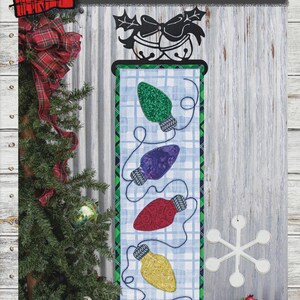 May include: A holiday-themed wall hanging with the text "LIGHT UP THE HOLIDAYS" and dimensions of 6" x 20". It features colorful, embroidered Christmas light bulbs on a blue plaid background. The design includes a black metal bell and bow accent.