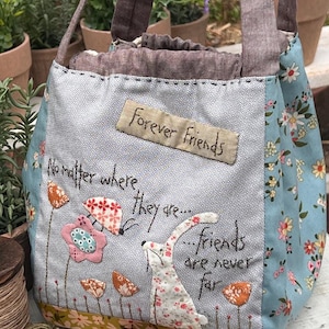 May include: A blue and white fabric tote bag with a floral pattern and the text "Forever Friends" and "No matter where they are... friends are never far". The bag has a brown fabric handle and a white bunny with a pink flower.