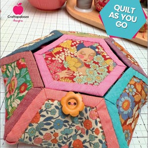May include: A Blossie Basket, a quilted fabric container with a floral design and a flower-shaped button closure. The basket features a variety of colorful fabrics, including pink, blue, and orange. The text "Quilt As You Go" is visible.