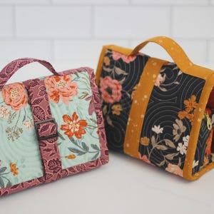 May include: Two quilted fabric bags with handles. One bag features a floral pattern in shades of pink, orange, and teal, with a buckle closure. The other bag has a black background with a floral design and mustard yellow accents.