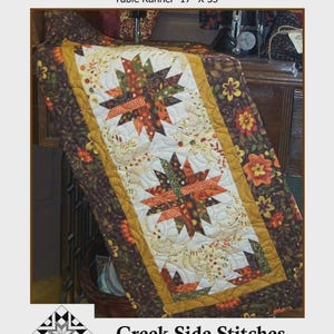 May include: A quilted table runner with a starburst design, featuring autumnal colors of orange, green, and brown. The runner is 17 inches by 35 inches, with the text "Charming Pineapple Star" and "Creek Side Stitches" visible.
