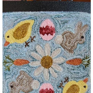 May include: A blue and white rug with an embroidered design of a daisy, two chicks, a bunny, and two eggs. The rug has the text "Garden Gals April" at the bottom.