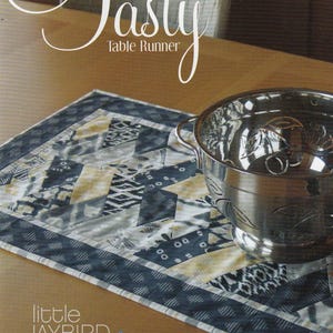 May include: A quilted table runner with a geometric pattern in shades of blue, gray, and yellow. The words "Tasty Table Runner" are written in a cursive font. A silver bowl sits on the runner.