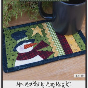May include: A rectangular fabric mug rug featuring a snowman appliqué with a black top hat, orange nose, and red scarf. The design includes a yellow star, green polka dot fabric, and a wavy gold trim. Text reads: Mr. McChilly Mug Rug Kit, 10" x 6".