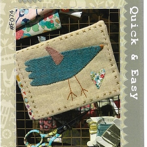 May include: A beige needlebook with a blue bird and a small floral heart design. The needlebook is on a wire rack with fabric scraps, scissors, and embroidery floss. The text "Hatched and Patched" is at the top of the image. The text "Quick & Easy Little Bird Needlebook" is at the bottom of the image. The text "#F074" is on the left side of the image.