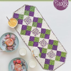 May include: A quilted table runner titled Fun Four-Patch Runner designed by Cathey Laird. The runner features a repeating pattern of purple, green, and white floral fabric squares. The text 14" x 35 1/2" is printed at the bottom of the image.