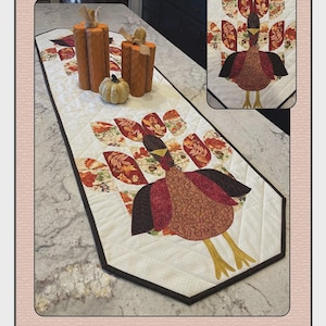 May include: A quilted table runner with a turkey design, featuring autumn colors. The runner has a white background with a black border. The image also includes wooden pumpkin decorations and the text "Turkey Thyme" and "Quilted Garden Designs".