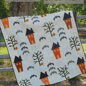 May include: A cream-colored quilt featuring a repeating pattern of orange houses with black roofs, dark green bare trees, small orange pumpkins, and grey flying bats. The fabric has a subtle quilted texture throughout the entire surface.