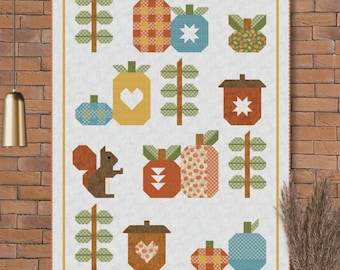 Puede incluir: Un edredón de retazos con un fondo crema presenta varios diseños con temática otoñal, incluyendo calabazas, bellotas y una ardilla. El edredón tiene un borde y el texto "Acorn Row" en la parte inferior. El edredón se exhibe en una pared de ladrillo.