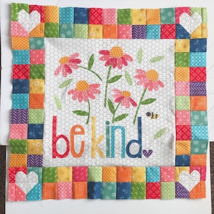 May include: A square fabric quilt block featuring a white background with a gray honeycomb pattern. The center displays four pink flowers with yellow centers, green leaves, a small bee, and the text be kind in colorful letters. A border of small multicolored fabric squares surrounds the center, with white heart shapes at each corner.