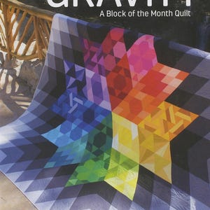 May include: A colorful quilt with a starburst design, featuring a gradient of colors from blue to red and yellow. The quilt is labeled "GRAVITY" and "A Block of the Month Quilt". The quilt is made by Jaybird Quilts.
