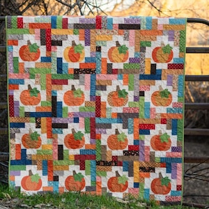 May include: A patchwork quilt with a white background and colorful squares featuring a pattern of pumpkins. The quilt is hanging on a metal gate.