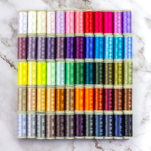 May include: A collection of 48 spools of thread in various colors, arranged in rows on a marble surface. The spools are all the same size and shape, and they are arranged in a rainbow pattern.