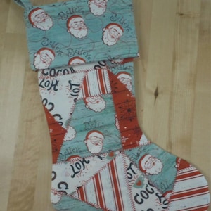 May include: A Christmas stocking featuring a patchwork design with alternating fabric patterns. The cuff is light blue with Santa Claus faces and the word Believe. The body includes red and white stripes, Santa faces, and text reading Hot Cocoa.